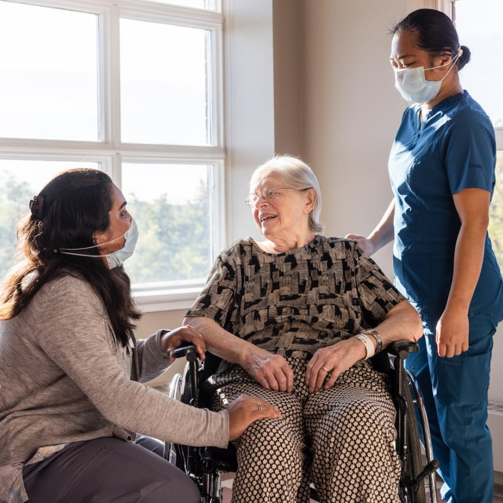 Two healthcare workers assist an elderly woman in a wheelchair.