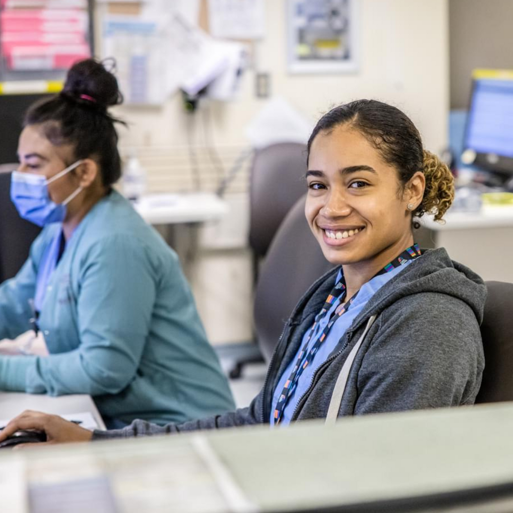 A healthcare worker with her hair tied back in a ponytail sits at her desk and smiles for the photo