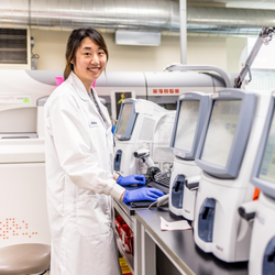 Image of A healthcare worker wearing a white lab coat and blue gloves stands in front of a computer in the Hospital's laboratory and smiles for the photo